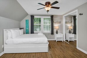 Een slaapkamer met een bed, een plafondventilator, een raam met zonwering, een nachtkastje met een lamp en een spiegel.