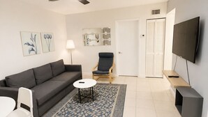 A modern living room with a grey sofa, a small round coffee table, a white chair, a floor lamp, a ceiling fan, a TV mounted on the wall, and a door with louvered shutters.