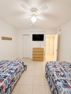 Un dormitorio con dos camas, ventilador de techo, televisor, cómoda y una puerta que da a otra habitación.