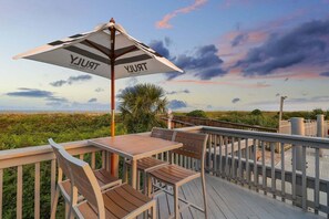 Eine Holzterrasse mit Tisch, Stühlen und Sonnenschirm, Blick auf Strand und Himmel.