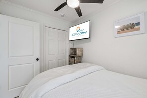 Une chambre avec un lit, un ventilateur de plafond, un téléviseur fixé au mur qui affiche « HOST & HOME, locations de vacances de luxe », et un tableau encadré accroché au mur.