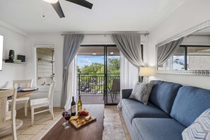 A living room with a sofa, a dining table with chairs, a balcony with a view of the street, and a small kitchen area. - Ocean Views | Community Pool, 2 BR, Sleeps 6, Walkable to Coligny Plaza! (Hilton Head Island)