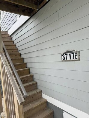 Un escalier en bois mène à une maison dont l’extérieur est recouvert de panneaux blancs. On peut voir une plaque avec le numéro de la maison sur le mur.