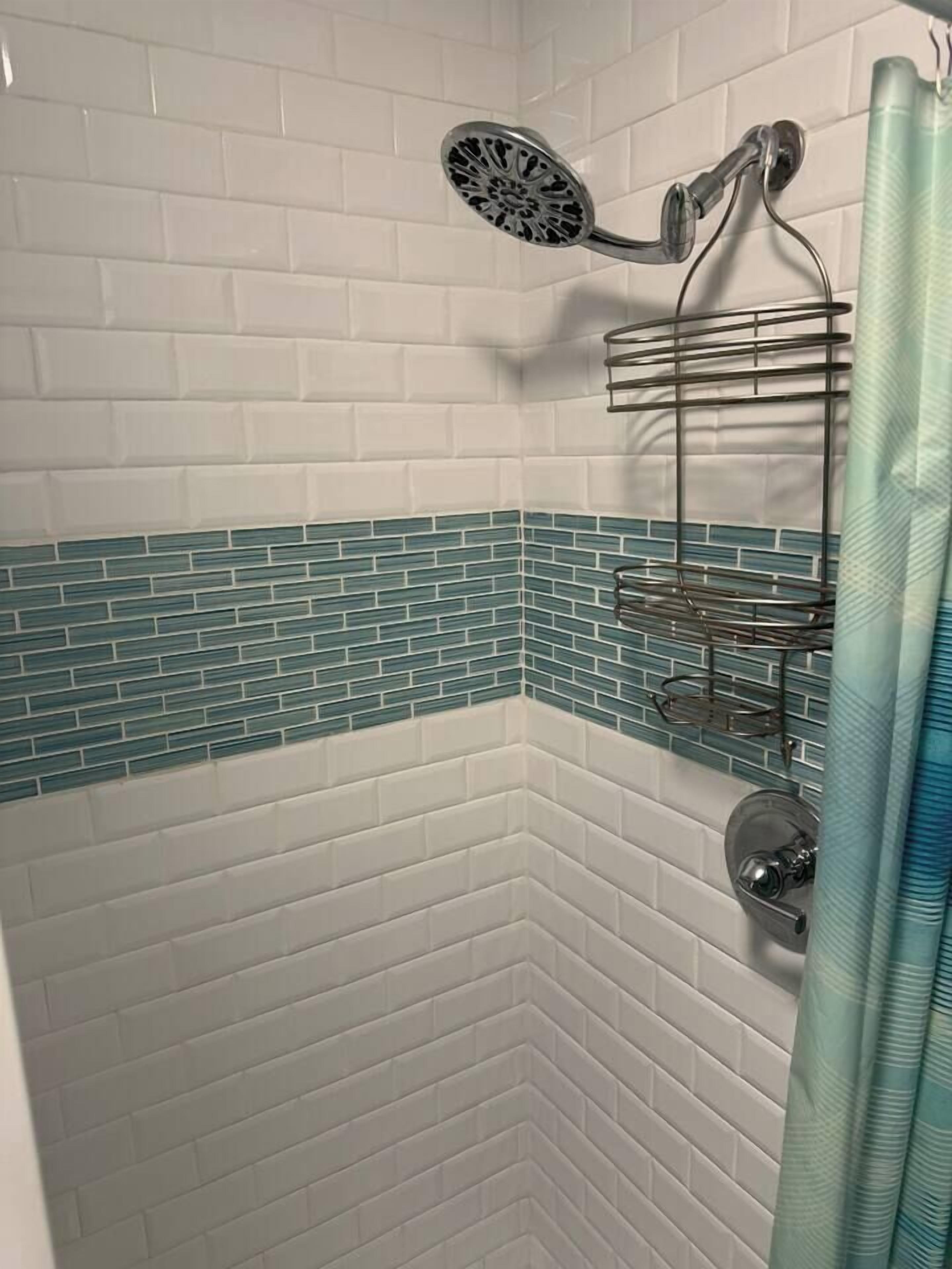 Une douche équipée d’un pommeau de douche à main, d’une étagère en métal et d’un mur carrelé avec un motif blanc et turquoise.