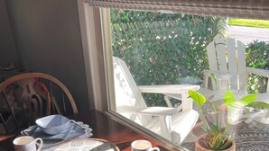 Mesa de comedor servida con platos, tazones y tazas, con vista a mobiliario exterior y follaje a través de una ventana.