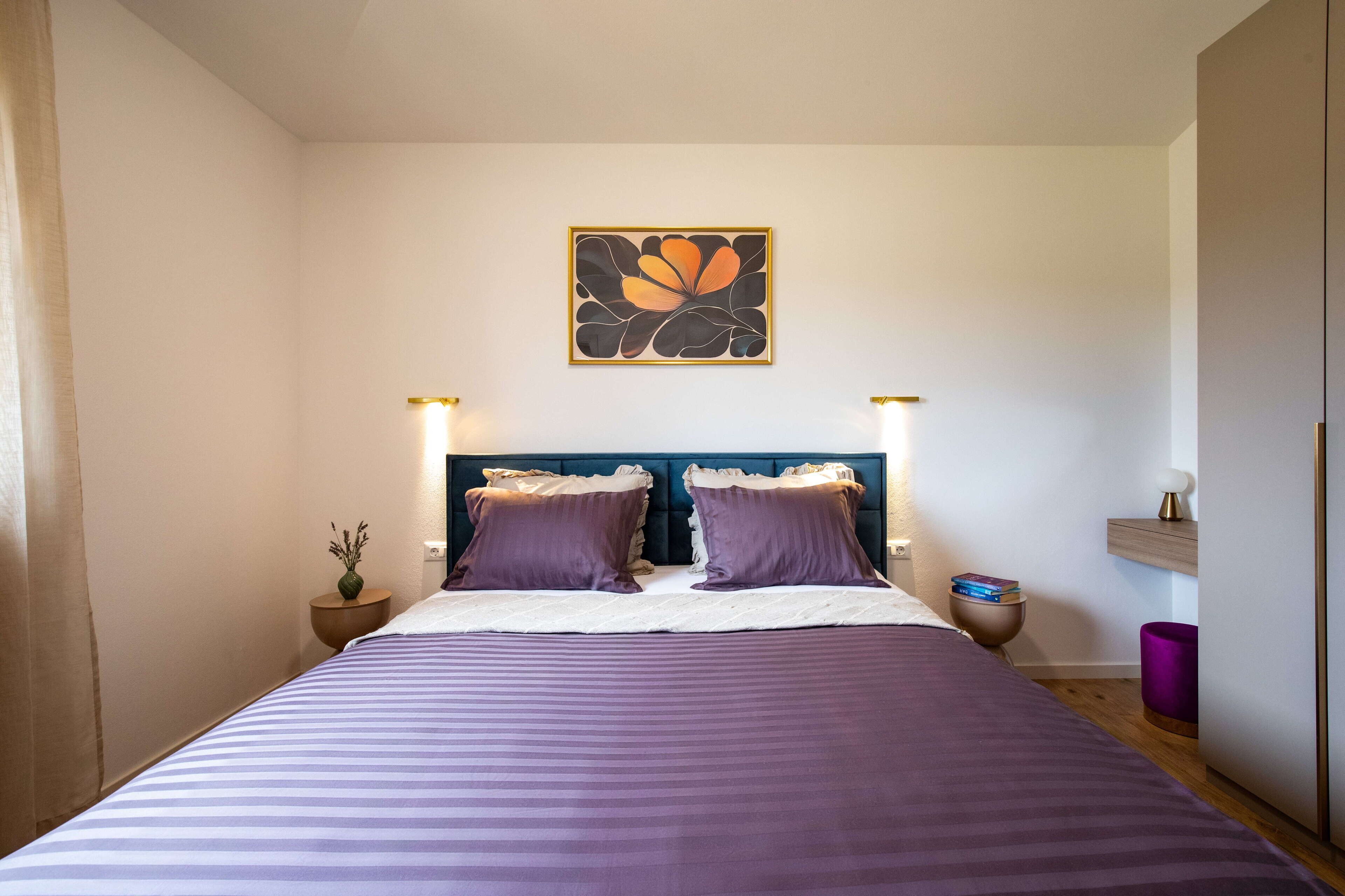 A bedroom with a bed, two bedside tables, a nightstand, a wall-mounted artwork, and a bedside lamp.