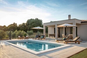 A rectangular swimming pool with lounge chairs and umbrellas, surrounded by a patio area with a modern building in the background. - Villa Casa Dvori (Istria - Valtura) (Valtura)