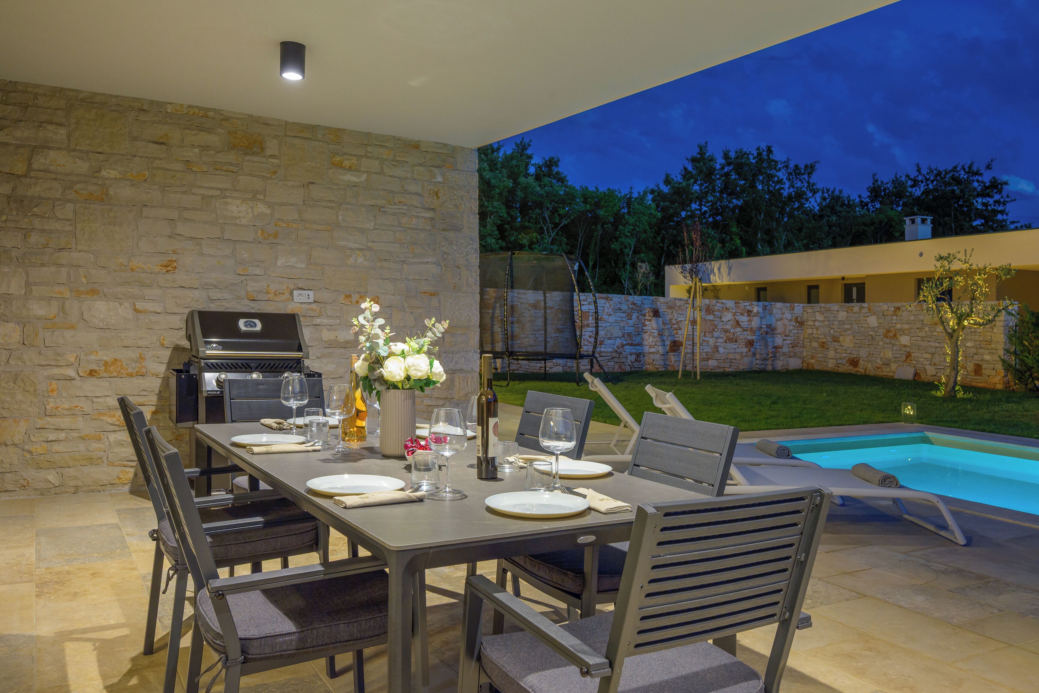 An outdoor dining area with a stone wall, a grill, and a pool in the background.