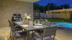 An outdoor dining area with a stone wall, a grill, and a pool in the background. - Villa Luen (Istria - Cukrići) (Cukrići)
