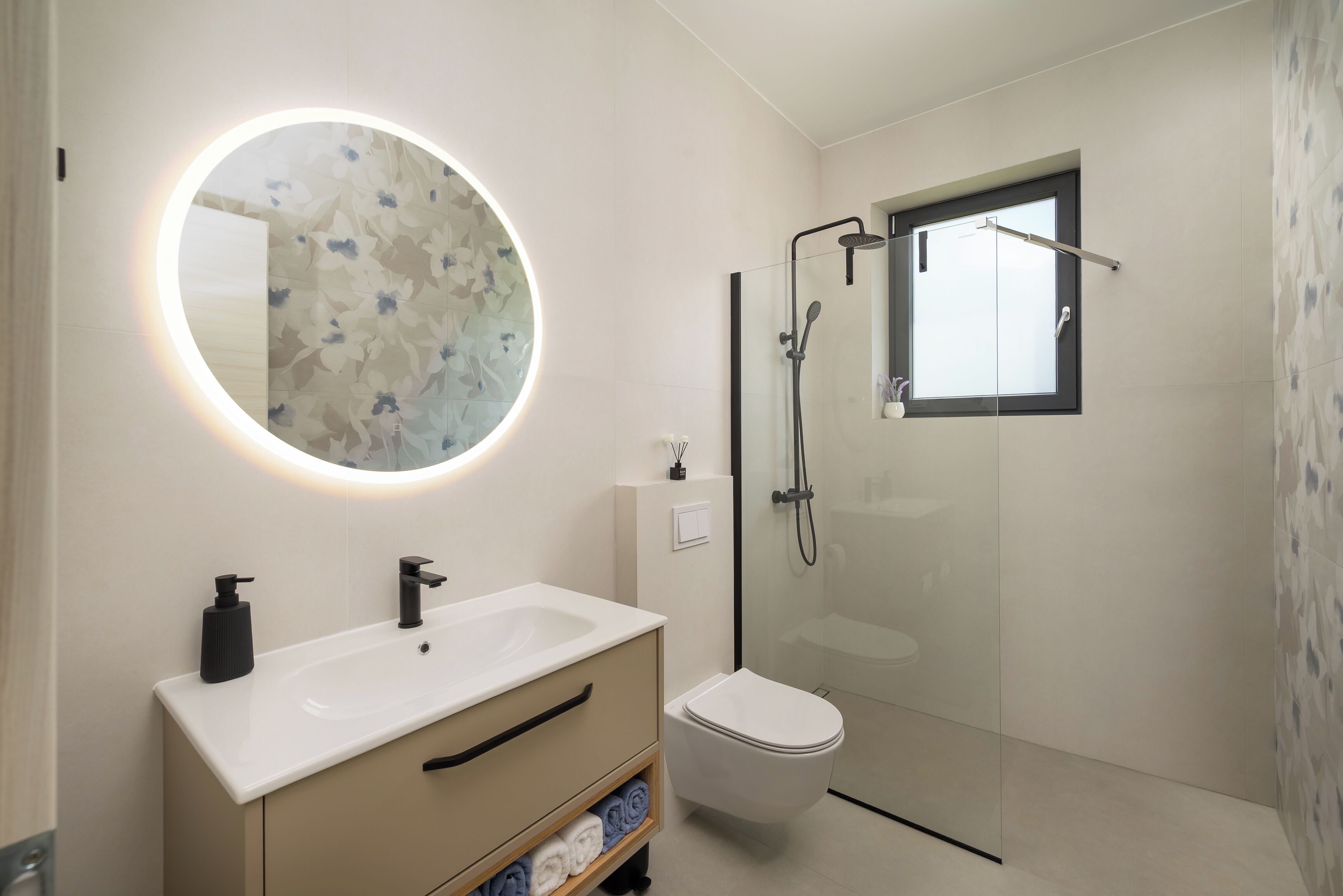 Ein modernes Badezimmer mit rundem Spiegel, Waschbecken in Weiß, Toilette und einer Duschkabine aus Glas.