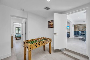 Ein modernes Interieur mit einem Kicker im Vordergrund, einem Essbereich mit blauer Couch und Tisch, und einem durchgegangene Tür erkennbaren Schlafzimmer.