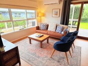 A living room with a sofa, a coffee table, and a chair. There is a window with a view of greenery outside. - Cosy Coastal Cottage in Red Rocks – Close to Beach & Phillip Island Attractions (Cowes)