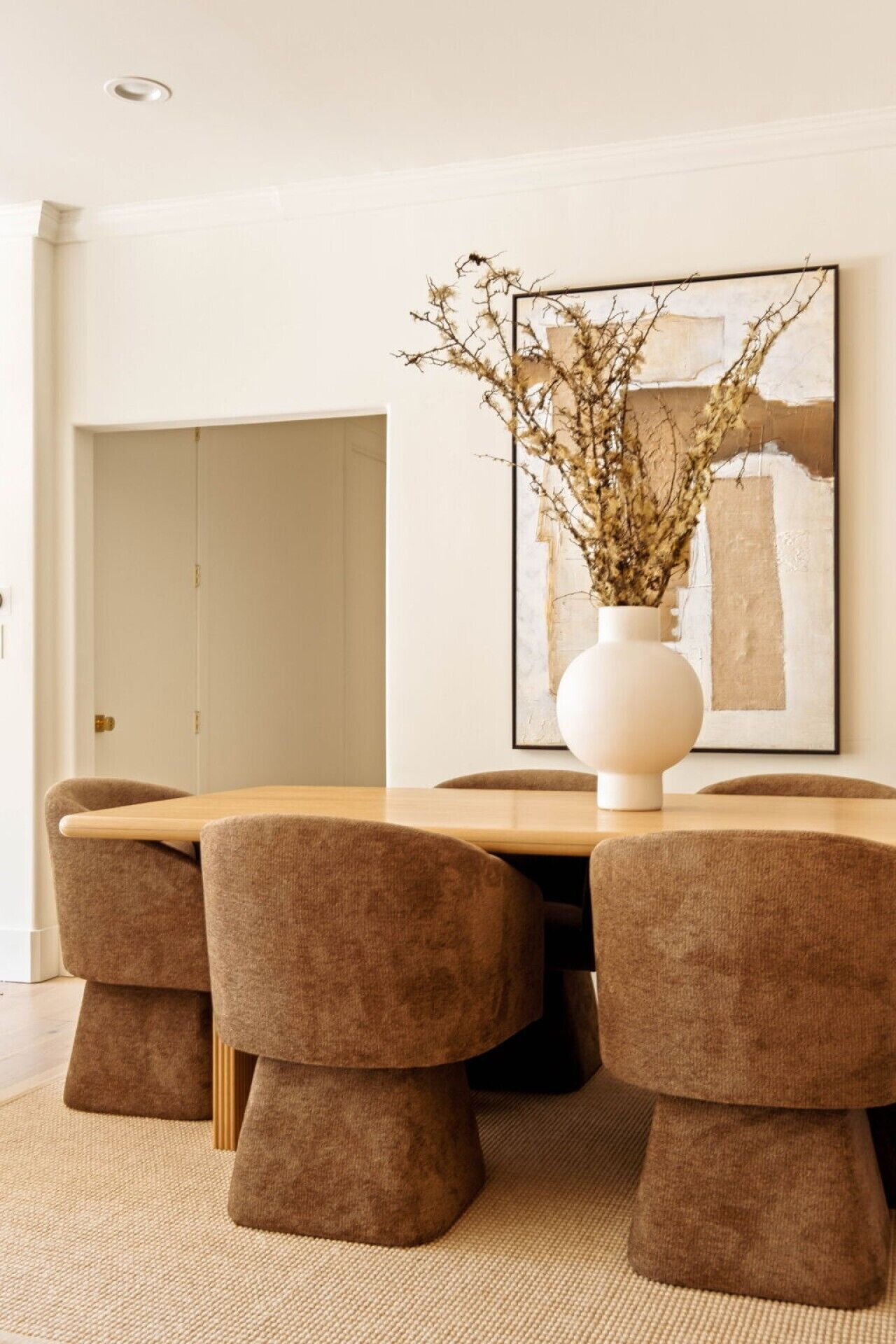 A dining area with a round table, four upholstered chairs, a white vase with branches, and a large abstract painting on the wall.