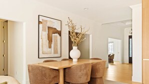 A dining area with a wooden table and cushioned chairs, a vase with dried flowers, and a large abstract painting on the wall.