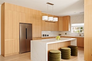 A modern kitchen with wooden cabinets, a stainless steel refrigerator, a marble countertop, and a hanging light fixture.