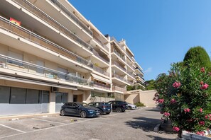 A multi-story residential building with balconies, a parking area with cars, and a well-maintained garden with flowering plants. - Appartement Les Heures Claires avec Terrasse - Welkeys (Mandelieu-la-Napoule)