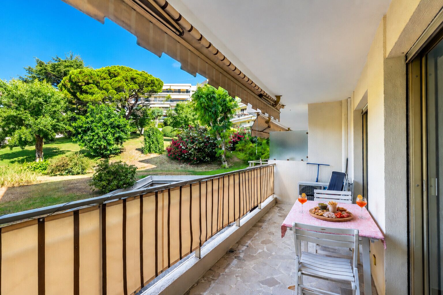 A balcony with a table set for two, overlooking a garden with various plants and a building in the background.