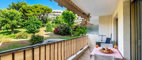 A balcony with a table set for two, overlooking a garden with various plants and a building in the background.