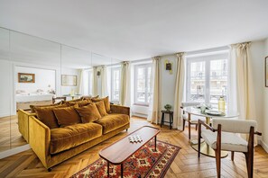Ein Wohnzimmer mit einer Couch, einem Couchtisch, einem Esstisch mit Stühlen und großen Fenstern mit Vorhängen.