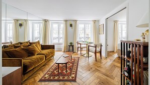 A living room with a brown sofa, a coffee table, a patterned rug, and a dining area with a table and chairs. - Le Nid Doré Parisien - Welkeys (Paris)