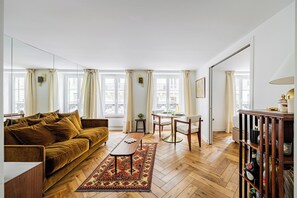 Ein Wohnzimmer mit braunem Sofa, einem Couchtisch, einem gemusterten Teppich und einem Essbereich mit Tisch und Stühlen.