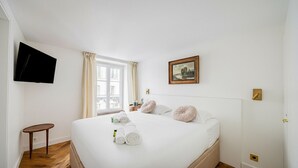 A hotel room with a bed, a TV mounted on the wall, a window with curtains, a bedside table, and a painting on the wall. - Le Nid Doré Parisien - Welkeys (Paris)