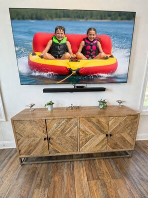 A television mounted on a wall displaying two children on a red inflatable raft. Below the TV, there's a wooden cabinet with a 'welcome' sign.