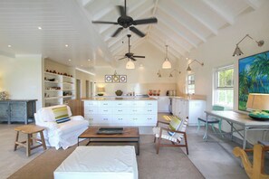A spacious living area with a white sofa, a coffee table, and a dining table with chairs. There is a kitchen area with white cabinets and a large window. - Cowrie Cottage | Private Pool | Near Gibbes Beach (Gibbes)