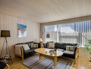 Sala de estar com sofá de couro preto, mesa de centro de madeira e uma grande janela com persianas.