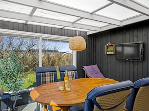 Uma sala de sol com mesa de madeira, cadeiras, televisão e vista para o exterior.