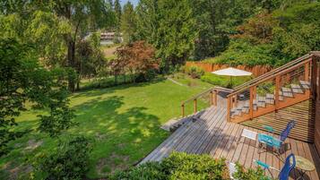 Um deck de madeira com móveis de exterior, gramado e vista para uma casa ao fundo.