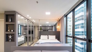 A modern bedroom with a large bed, a seating area, and a view of a cityscape through large windows. - Ap in Condo With Pool 350m From Meia Praia Ift0808 (Itapema)