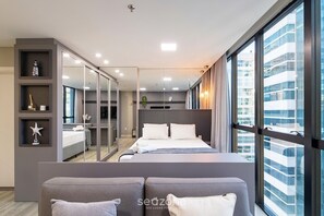 A modern bedroom with a large bed, a seating area, and a view of a cityscape through large windows. - Ap in Condo With Pool 350m From Meia Praia Ift0808 (Itapema)