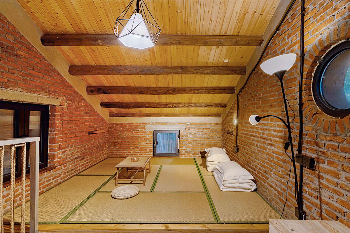 A room with a brick wall, wooden beams, tatami mats, a small table, a lamp, and a window with a curtain.