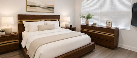 A bedroom with a large bed, wooden headboard, bedside tables with lamps, a nightstand, a dresser, a window with blinds, and a framed picture on the dresser.