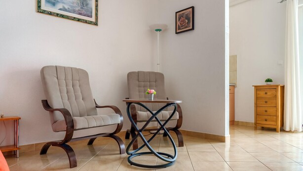 Ein Zimmer mit zwei Sesseln, einem kleinen Tisch mit einer Vase, einer Lampe und einem Holzschrank.