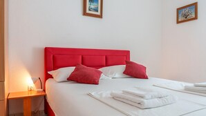 Uma cama bem arrumada com lençóis brancos e almofadas vermelhas, uma mesa de cabeceira com abajur e duas quadros pendurados na parede.