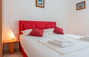 Ein ordentlich bezogenes Bett mit weißen Leinentüchern und roten Kissen, ein Nachttisch mit Lampe sowie zwei gerahmte Bilder an der Wand.
