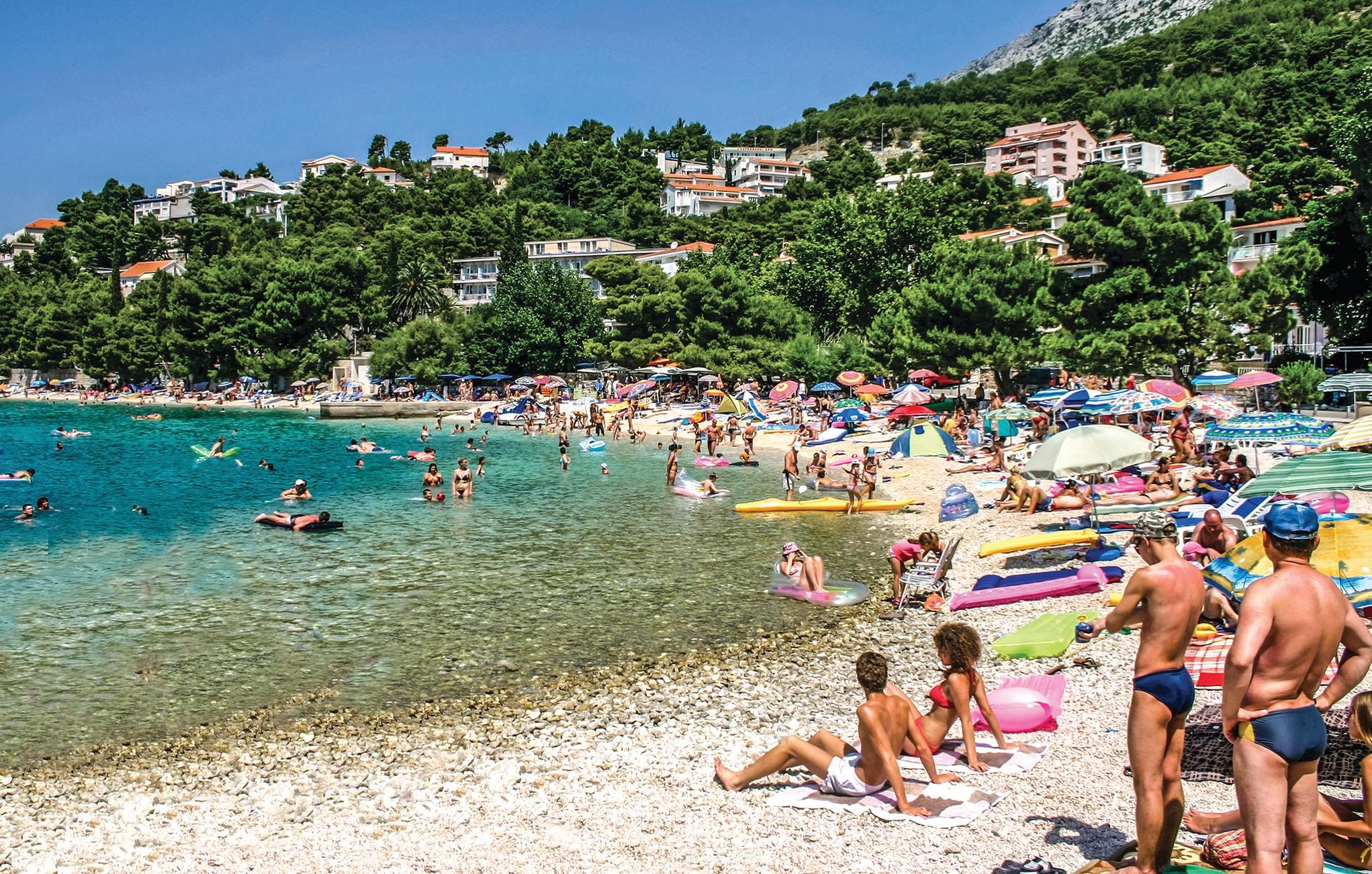 A crowded beach with clear turquoise water, people swimming and sunbathing, and buildings in the background.