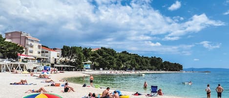 A beach with people sunbathing and swimming, a colorful umbrella, and buildings in the background.