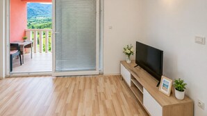 Una habitación con piso de madera, un televisor sobre un mueble de madera, una puerta corrediza de vidrio a un balcón y una pequeña mesa con una silla.