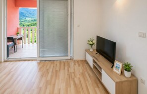 Ein Zimmer mit Holzboden, einem Fernsehgerät auf einem Holzschrank, einer Schiebetür zu einem Balkon und einem kleinen Tisch mit einem Stuhl.