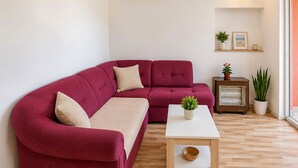 Una sala moderna con un sofá seccional rojo, una mesa de centro blanca y una mesita auxiliar con un calefactor. Hay plantas en macetas y un cuadro en un estante.