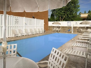 A swimming pool with white chairs and umbrellas, a lifeguard warning sign, and a brick wall in the background.