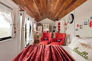 Un dormitorio con techo de madera, una cama con manta roja, dos sillas rojas, un escritorio y un reloj de pared.