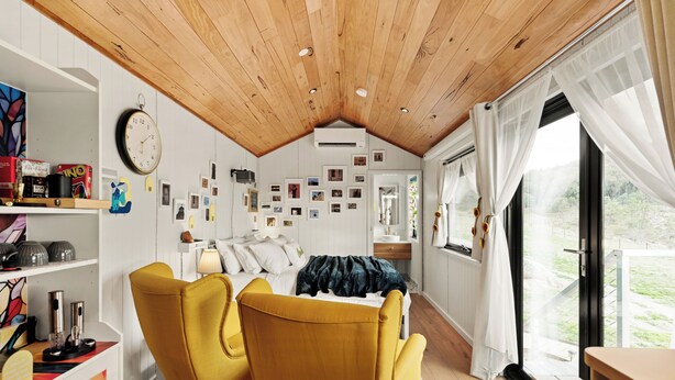 A cozy room with a bed, two yellow chairs, a wooden ceiling, and a large window with curtains.