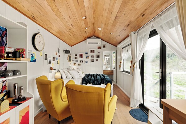 A cozy room with a bed, two yellow chairs, a wooden ceiling, and a large window with curtains.