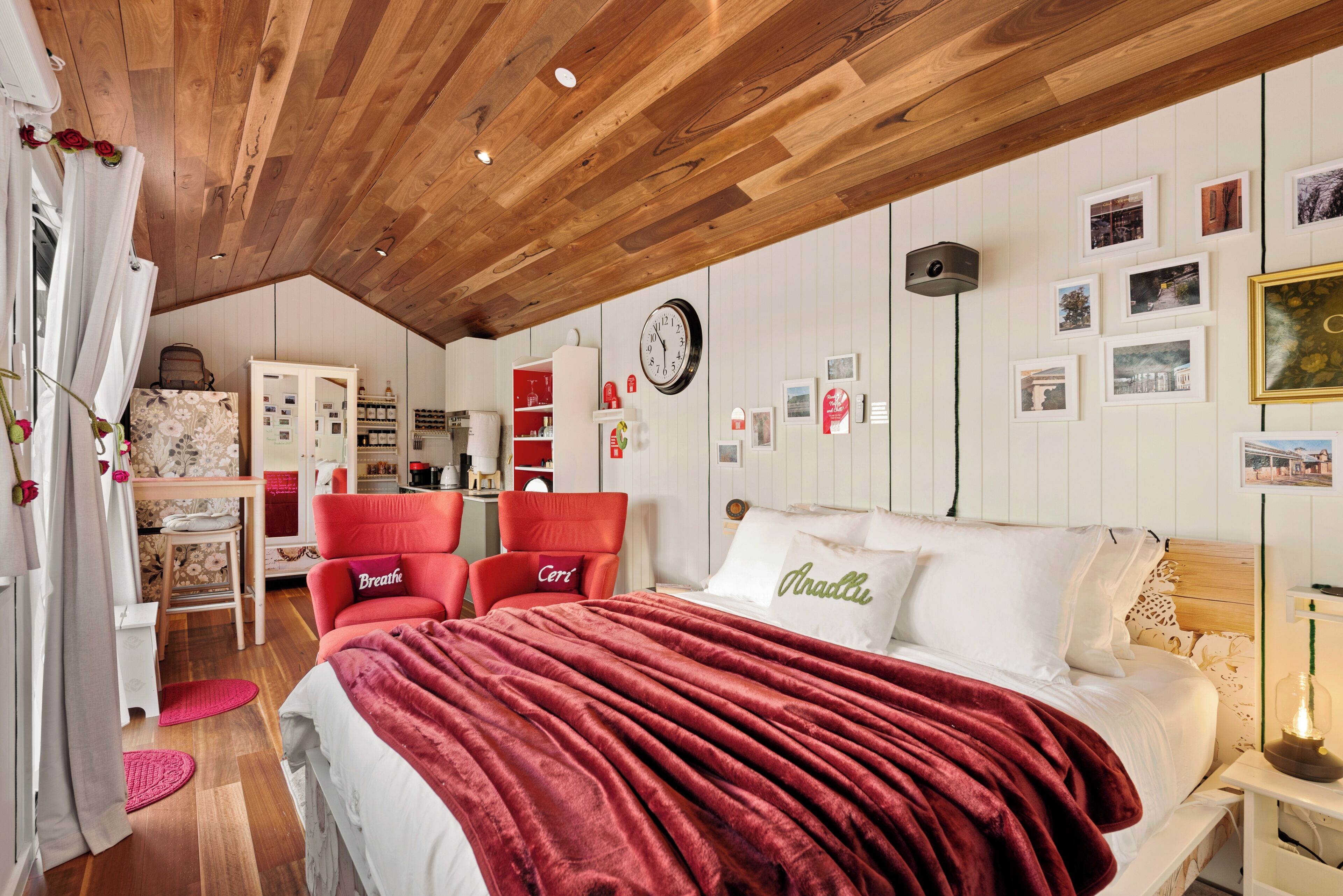 Un dormitorio con techo de madera, una cama con manta roja, dos sillas rojas, un armario blanco y un reloj de pared.