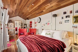 Une chambre avec un plafond en bois, un lit recouvert d’une couverture rouge, deux chaises rouges, une armoire blanche et une horloge murale.
