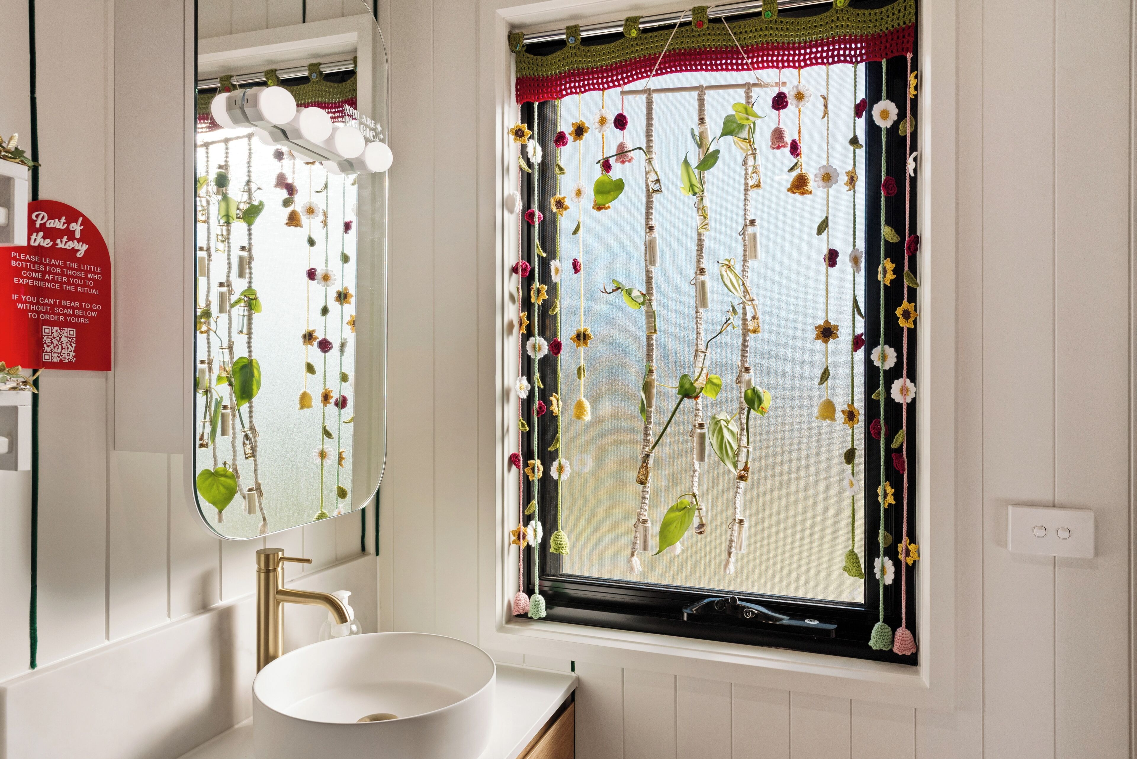 A bathroom with a white sink, a mirror, and a window with a crochet curtain.